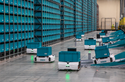 White mobile robots carrying teal totes in front of large storage system