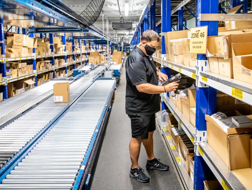 Man dressed in black scanning boxes in warehouse with conveyor behind him